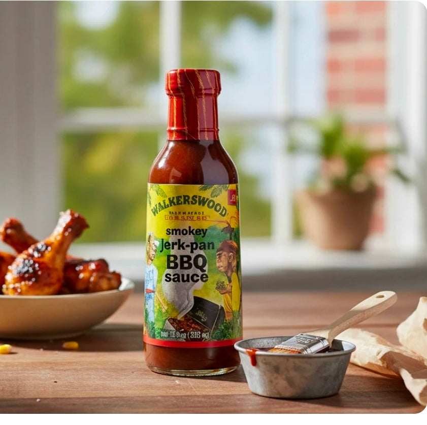 Bottle of Walkerswood smoky jerk pan BBQ sauce on a wooden table with food in the background